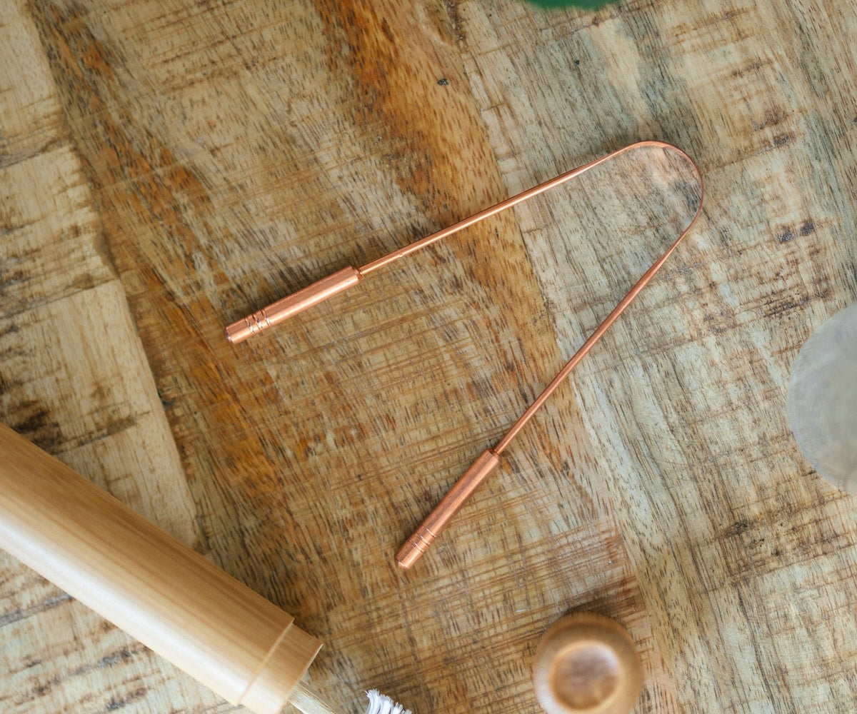Kopero Koperen Tongschraper, op een houten plank naast een bamboe tandenborstel