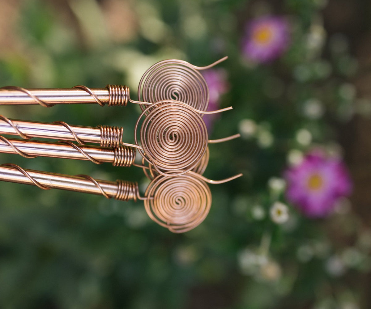 Close-up van koperen elektrocultuur-antennes met opgerolde spiraalkoppen, met bloemen op de achtergrond