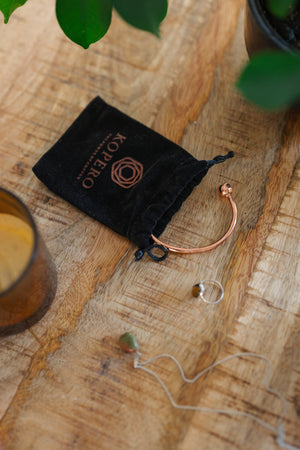 Kopero Koperen Armband Rond, op een houten tafel naast de fluwelen draagzak, een kaars en sieraden