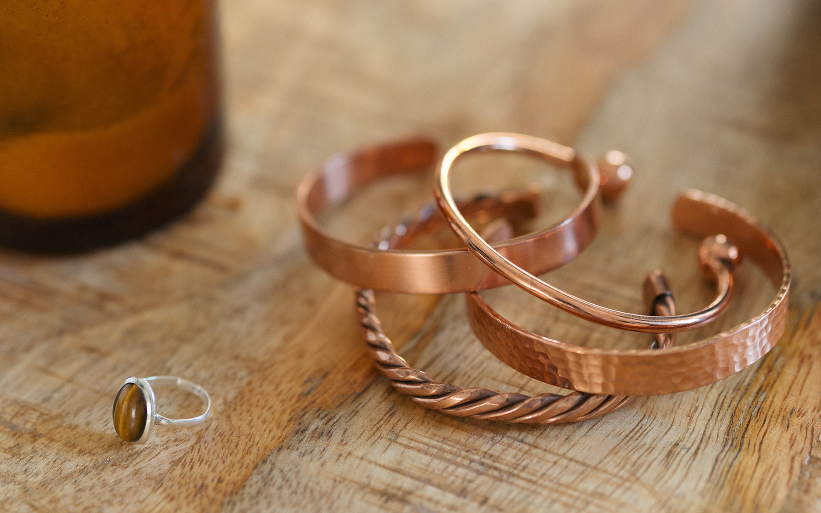 Vier koperen armbanden, gedraaid, glad, gehamerd en rond, op een houten tafel naast een kaars en een ring