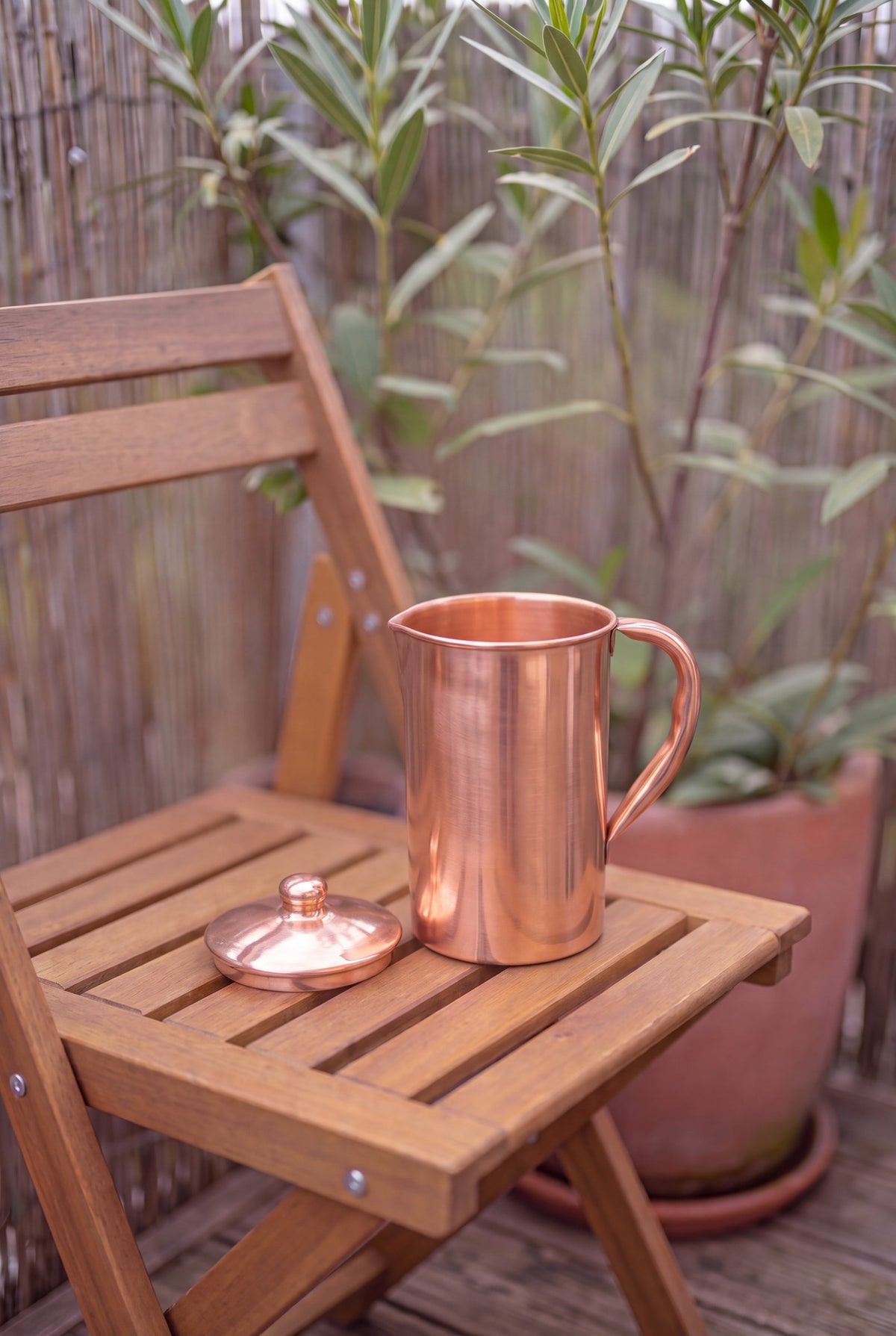 Cruche en cuivre Kopero Lisse en extérieur sur une chaise de jardin en bois, avec le couvercle à côté