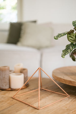 Kopero Koperen Piramide Energizer Gedraaid Groot (23,56 cm), op een houten salontafel voor een bank, naast kaarsen