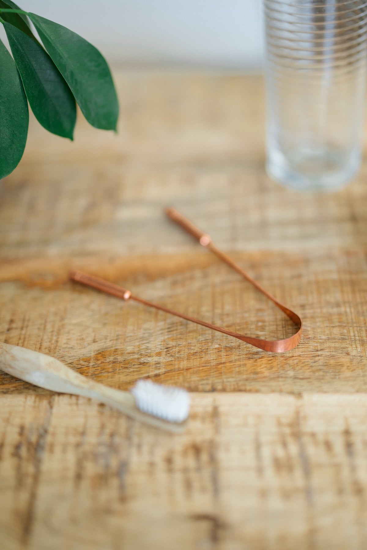 Kopero Koperen Tongschraper, op een houten tafel naast een bamboe tandenborstel en een glas water