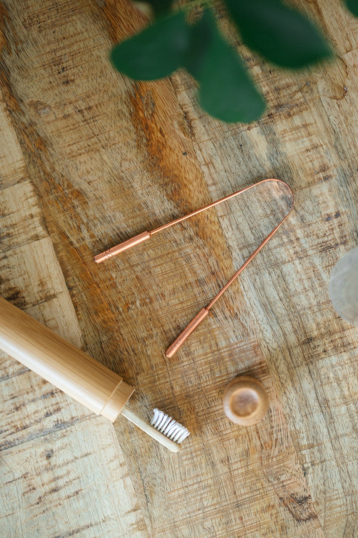Kopero Koperen Tongschraper, op een houten plank naast een bamboe tandenborstel