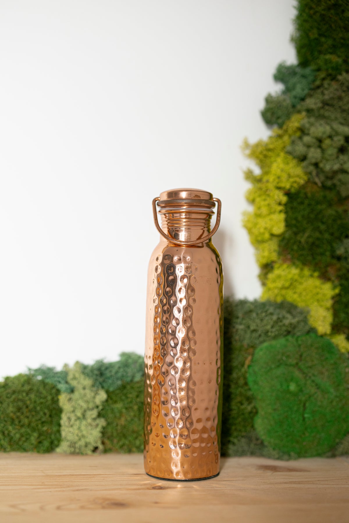 Gourde en cuivre Kopero Martelée, 900 ml, debout sur une table en bois devant un fond de mousse