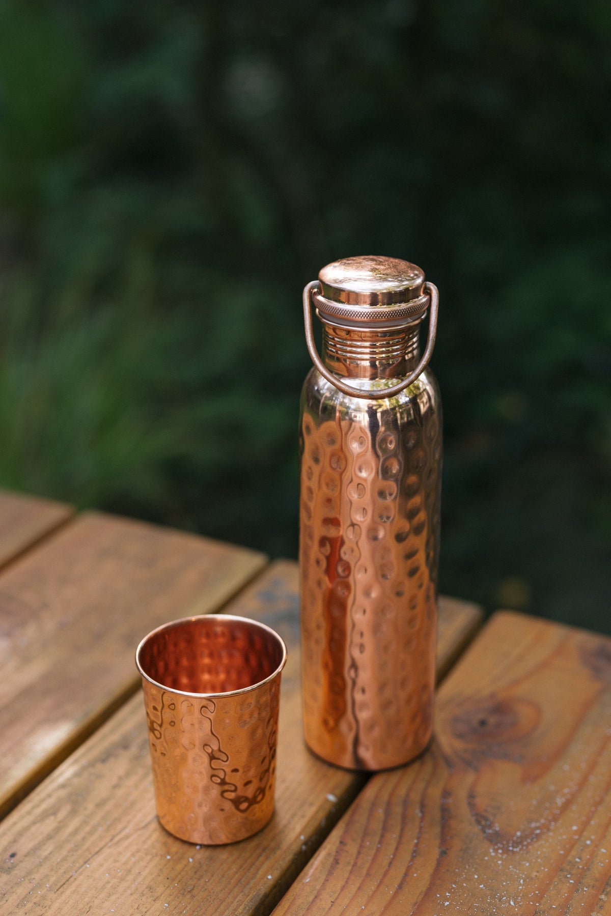 Kopero Koperen Waterfles Gehamerd, op een houten tuintafel naast een gehamerde beker