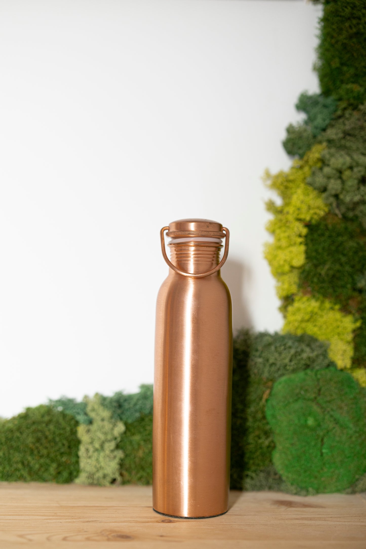 Gourde en cuivre Kopero Lisse, 900 ml, debout sur une table en bois devant un fond de mousse