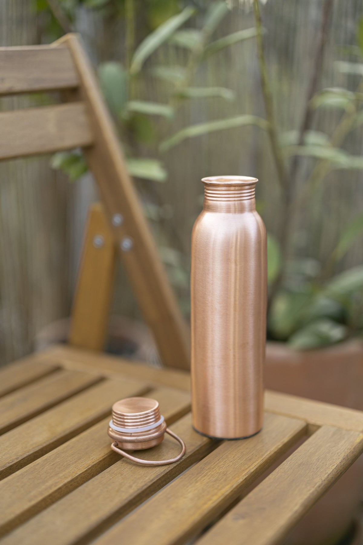 Bouteille d'eau en cuivre Kopero Lisse sur une chaise de jardin en bois, avec le bouchon à côté